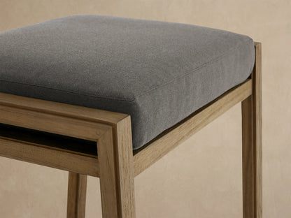 Canyon Outdoor Teak Backless Barstool with a light natural teak frame and dark grey cushion, against a beige wall & tiled floor.