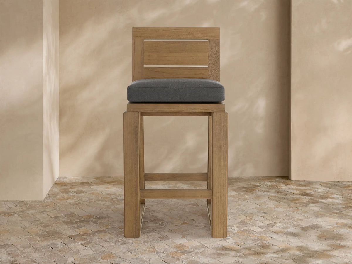 A Canyon Outdoor Teak Barstool. Light-toned teak wood with a slatted back and dark gray cushion, on a tiled patio.