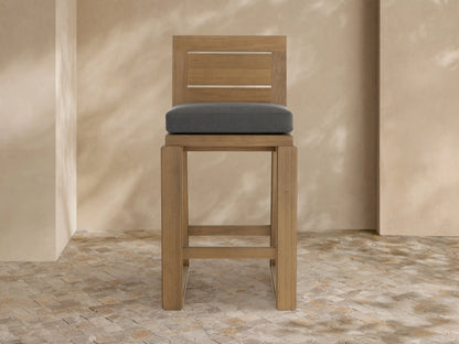 A Canyon Outdoor Teak Barstool. Light-toned teak wood with a slatted back and dark gray cushion, on a tiled patio.