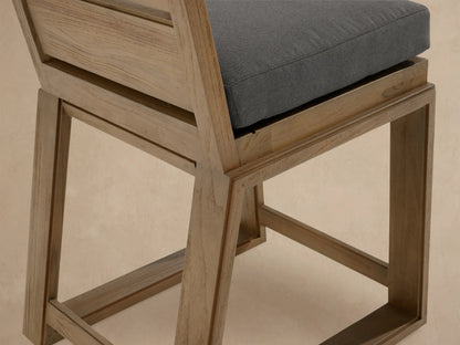 Canyon Outdoor Teak Barstool, light natural wood with a dark gray cushion, standing on a stone patio against a textured beige wall.