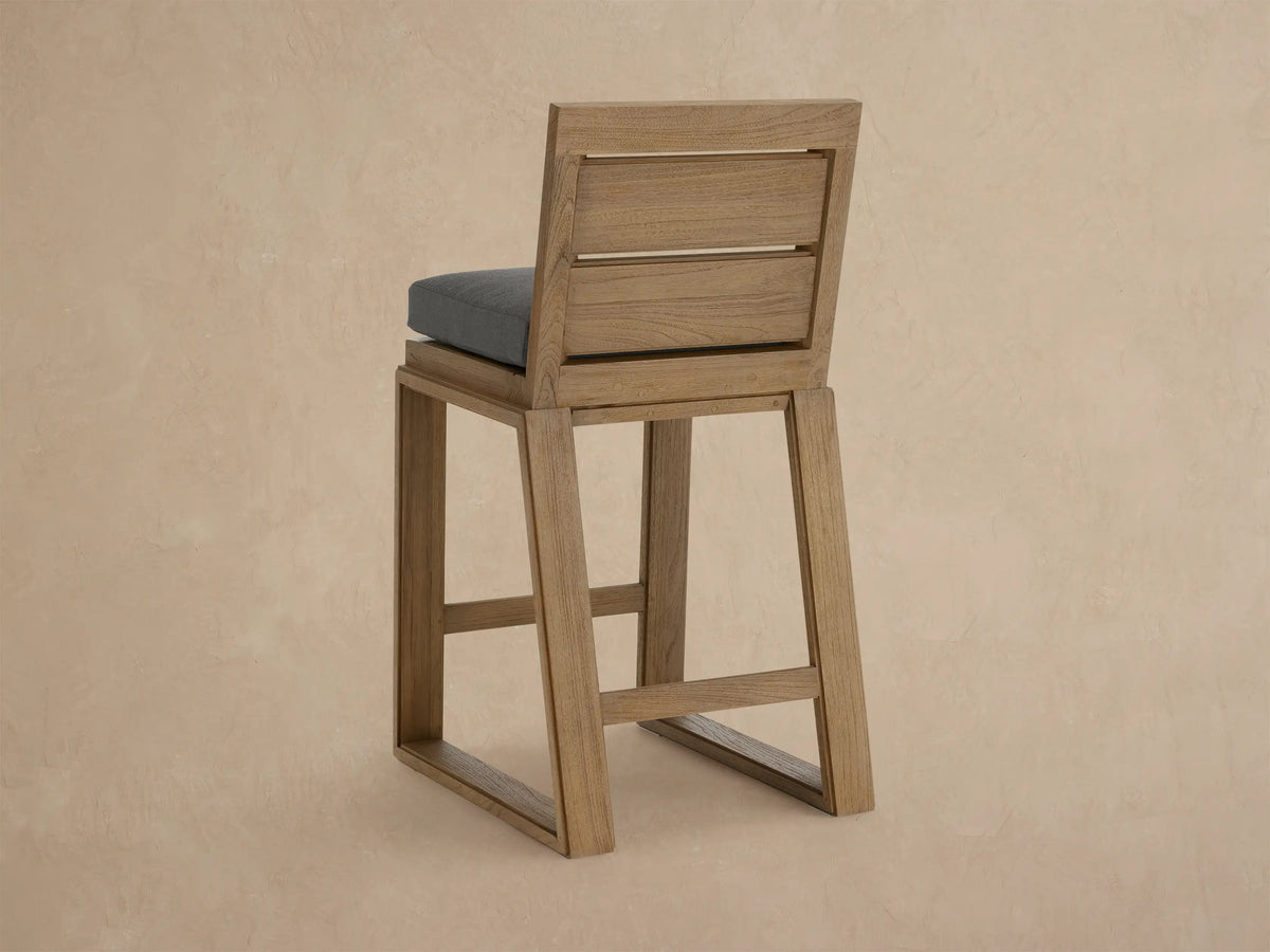 Angled close-up of a Canyon Outdoor Teak Barstool with a light brown, wire-brushed teak frame and a dark grey cushion.