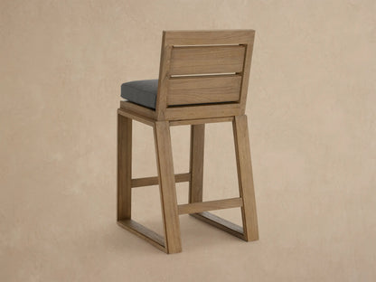 Angled close-up of a Canyon Outdoor Teak Barstool with a light brown, wire-brushed teak frame and a dark grey cushion.