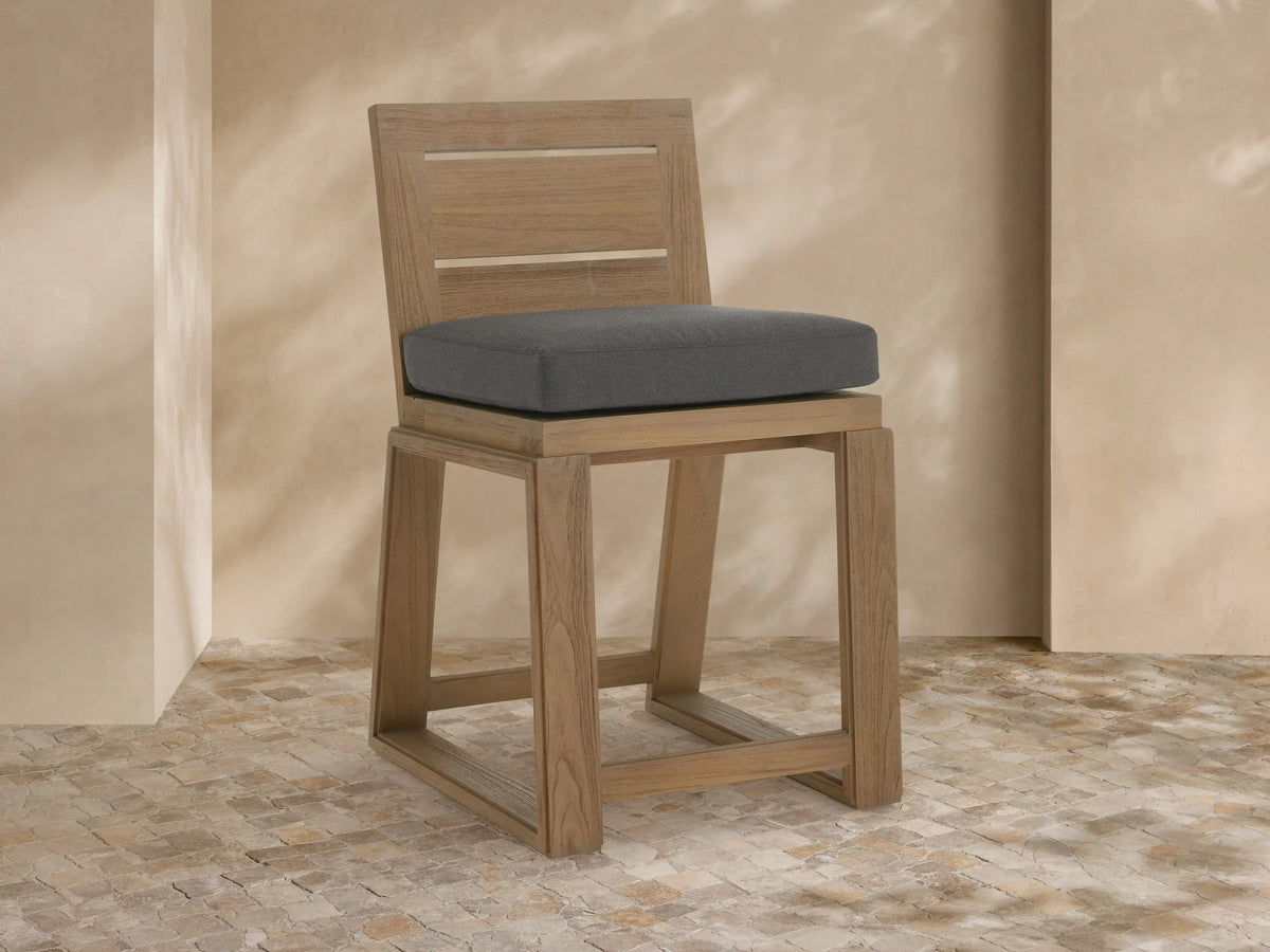 Canyon Outdoor Teak Counter Stool: light teak wood frame, dark grey cushion, angled right on stone floor, beige wall background.