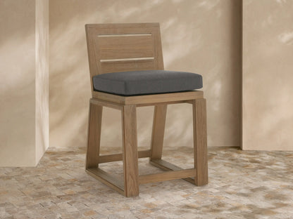 Canyon Outdoor Teak Counter Stool: light teak wood frame, dark grey cushion, angled right on stone floor, beige wall background.