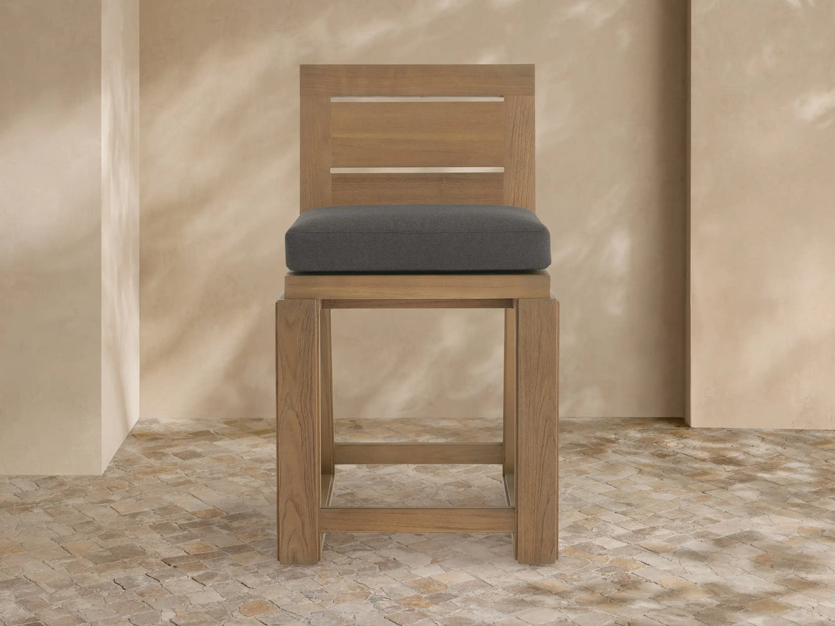 Canyon Outdoor Teak Counter Stool: light teak wood frame, dark grey cushion, angled right on stone floor, beige wall background.