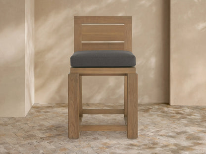 Canyon Outdoor Teak Counter Stool: light teak wood frame, dark grey cushion, angled right on stone floor, beige wall background.