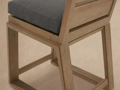 Canyon Outdoor Teak Counter Stool, light brown wood frame, slatted back, dark gray cushion, against a beige wall on tiled floor.