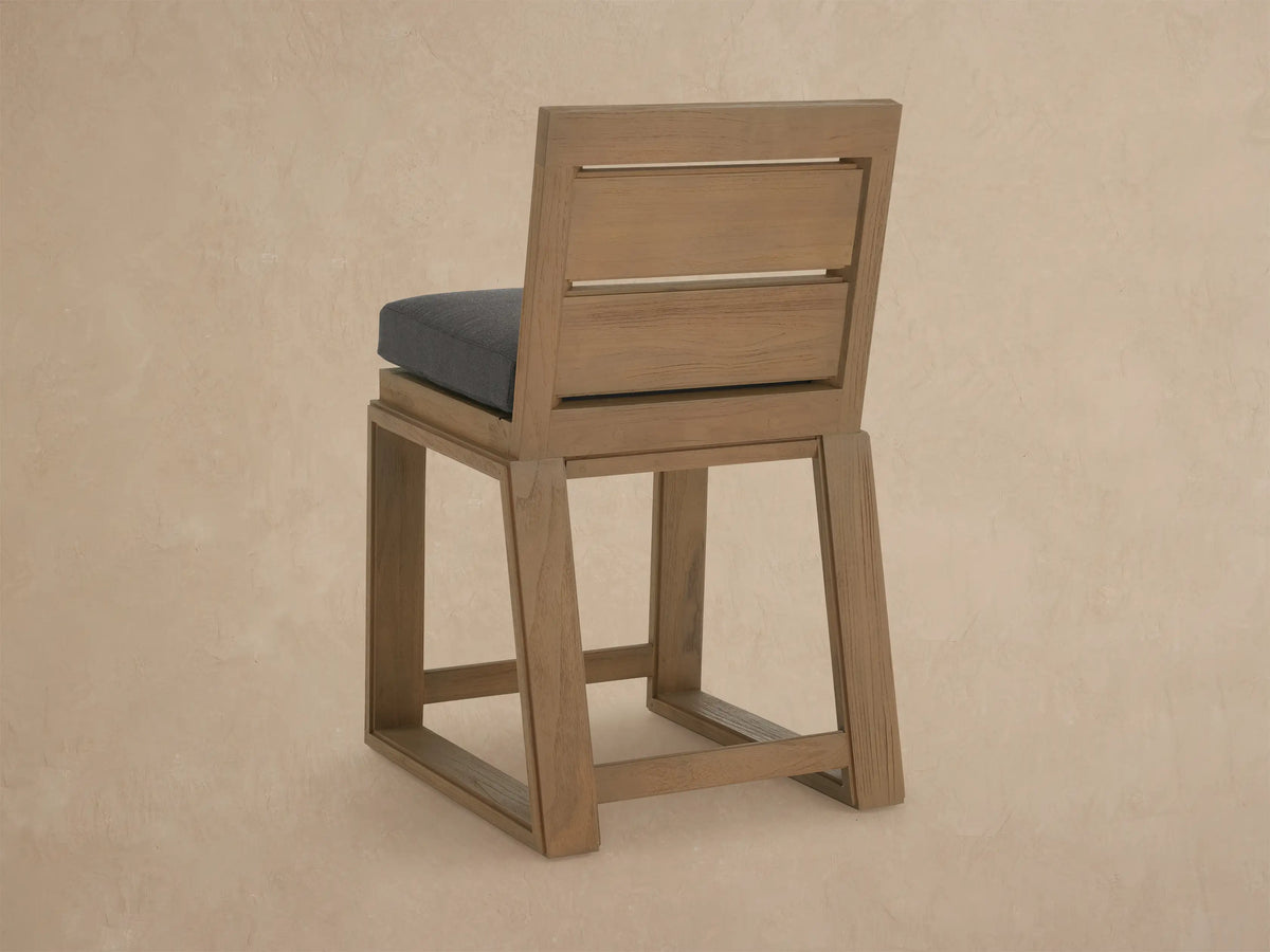 Angled close-up of a Canyon Outdoor Teak Counter Stool. Features light brown, distressed teak wood frame and dark gray cushion.