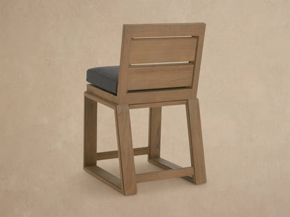 Angled close-up of a Canyon Outdoor Teak Counter Stool. Features light brown, distressed teak wood frame and dark gray cushion.