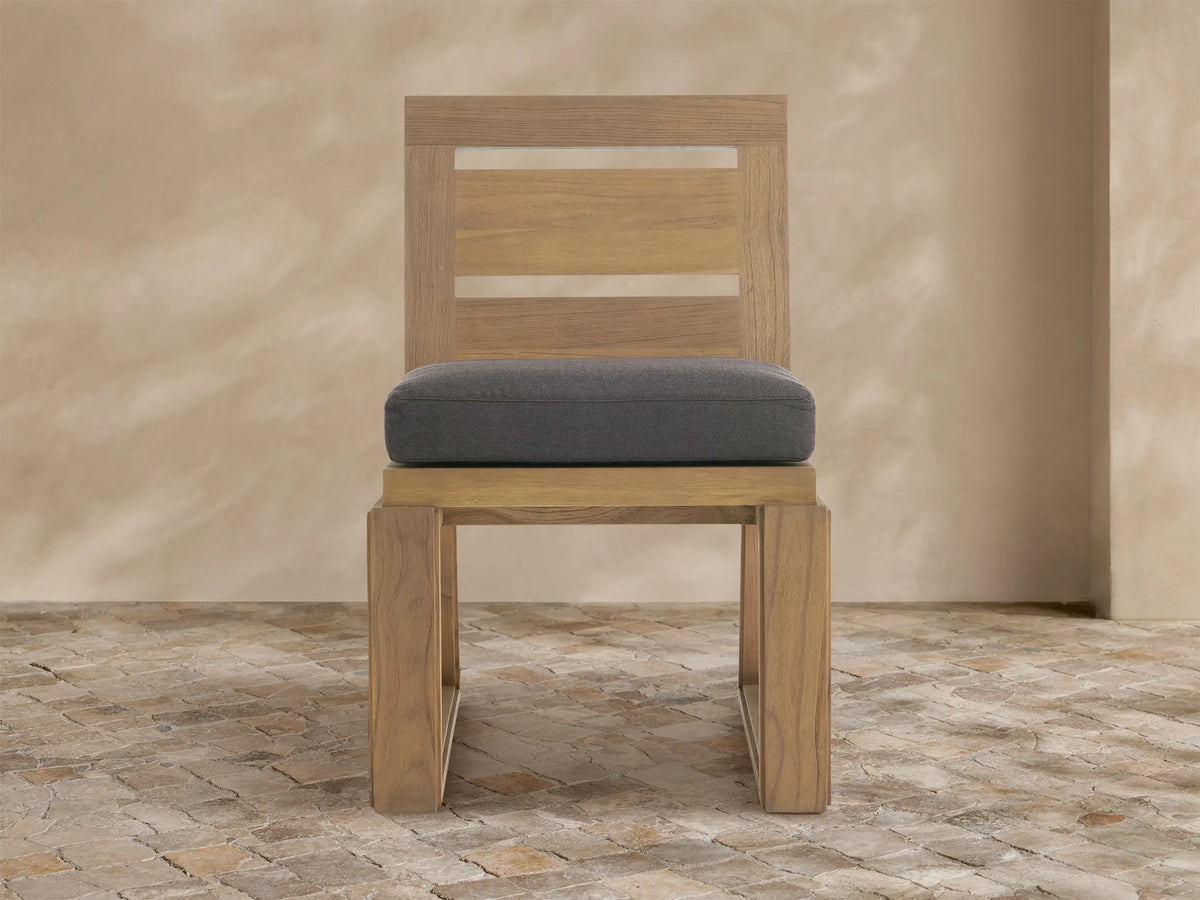 Canyon Outdoor Teak Dining Side Chair, light weathered teak with slatted back, dark gray cushion, on stone floor.
