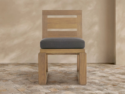 Canyon Outdoor Teak Dining Side Chair, light weathered teak with slatted back, dark gray cushion, on stone floor.