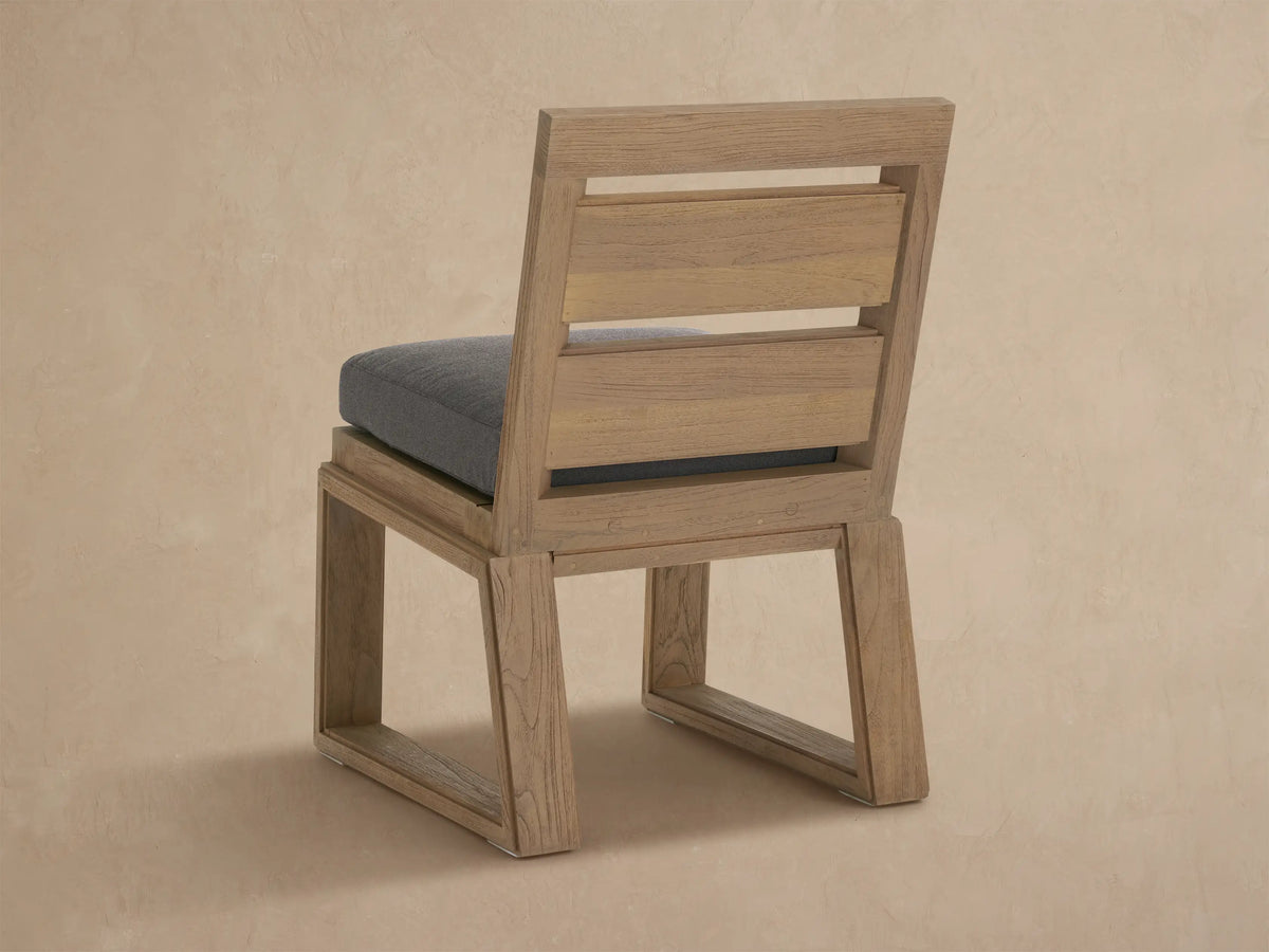 Angled close-up of Canyon Outdoor Teak Dining Side Chair. Light, textured teak wood frame holds a dark gray woven seat cushion.