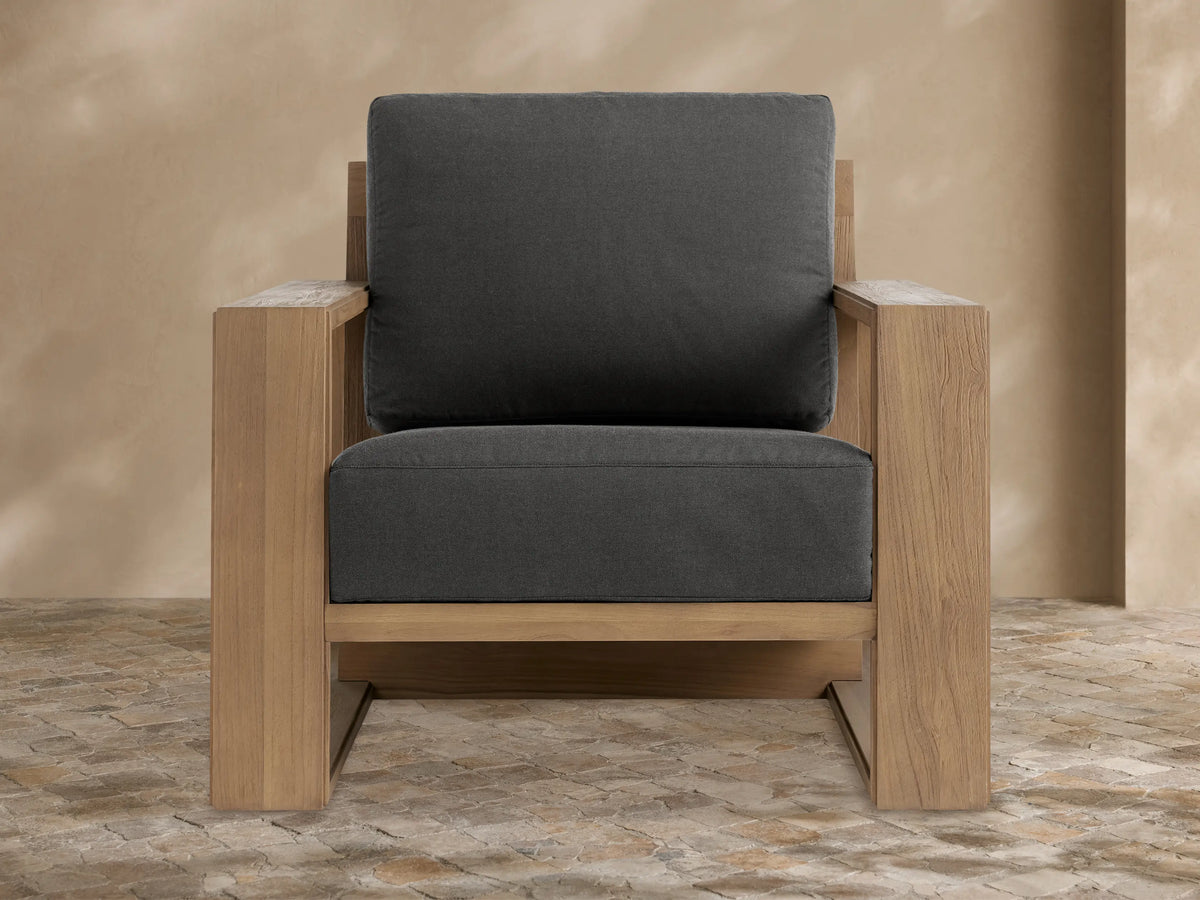 Canyon Outdoor Teak Lounge Chair with a light brown wood frame and dark gray cushions, angled right on a stone patio.