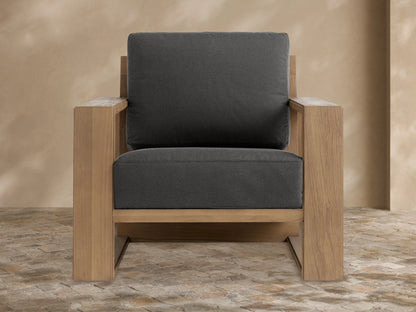 Canyon Outdoor Teak Lounge Chair with a light brown wood frame and dark gray cushions, angled right on a stone patio.
