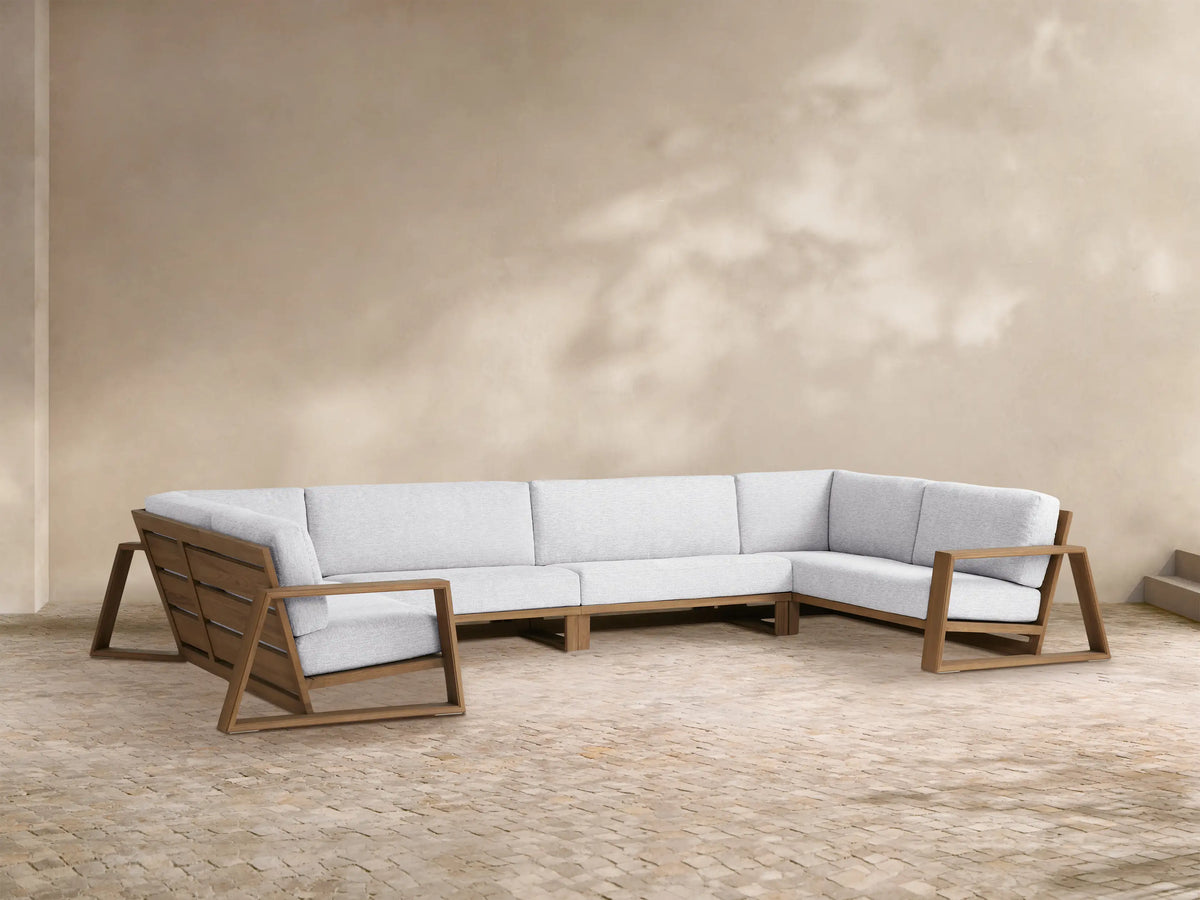 Canyon Outdoor Teak Four-Piece Sectional. L-shaped modular sofa with teak frames & light gray cushions, plus 2 armchairs on a patio.