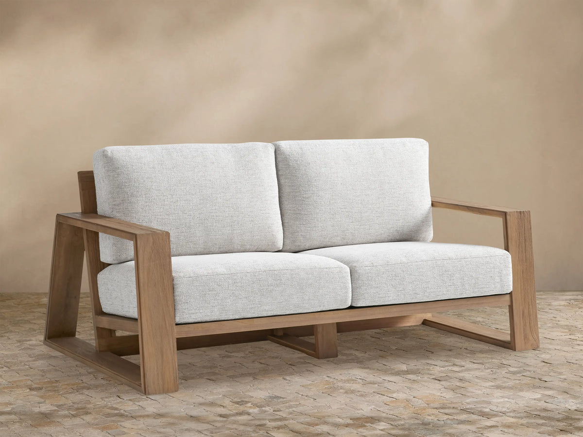 Canyon Outdoor Teak Sofa. Front view of a 2-seater with warm brown teak frame, light grey cushions, on a stone patio.