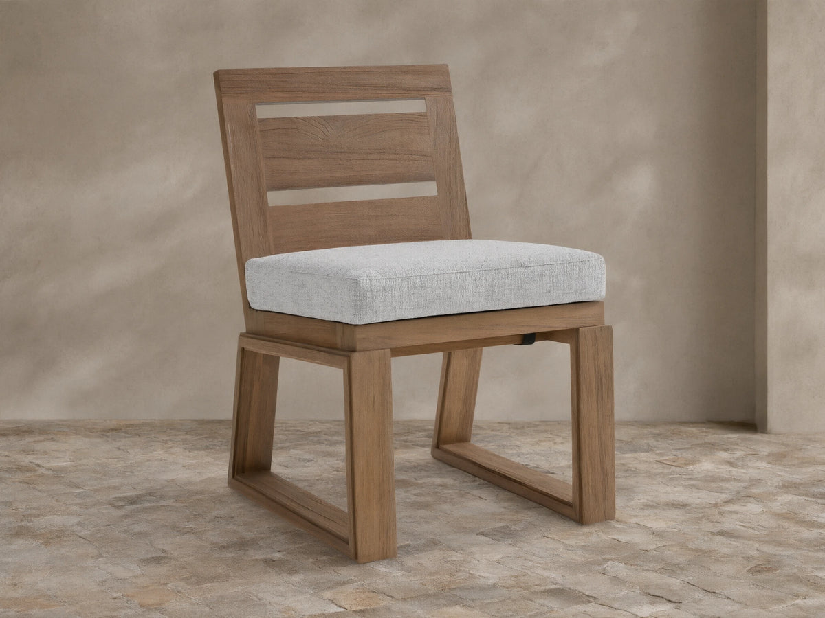 Canyon Outdoor Teak Dining Side Chair with light gray cushion & slatted back, on stone patio against beige wall.