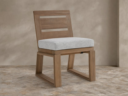 Canyon Outdoor Teak Dining Side Chair with light gray cushion & slatted back, on stone patio against beige wall.