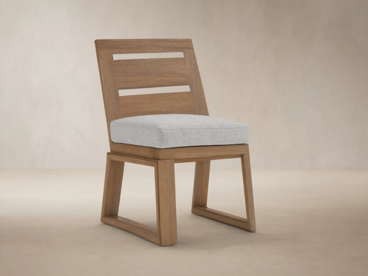 Canyon Outdoor Teak Dining Side Chair with light gray cushion & slatted back, on stone patio against beige wall.