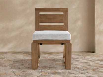 Canyon Outdoor Teak Dining Side Chair with light gray cushion & slatted back, on stone patio against beige wall.