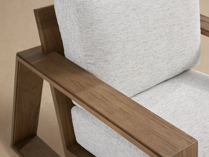 Front view of Canyon Outdoor Teak Lounge Chair with a brown teak frame and light grey cushions, on a stone floor against a beige wall.