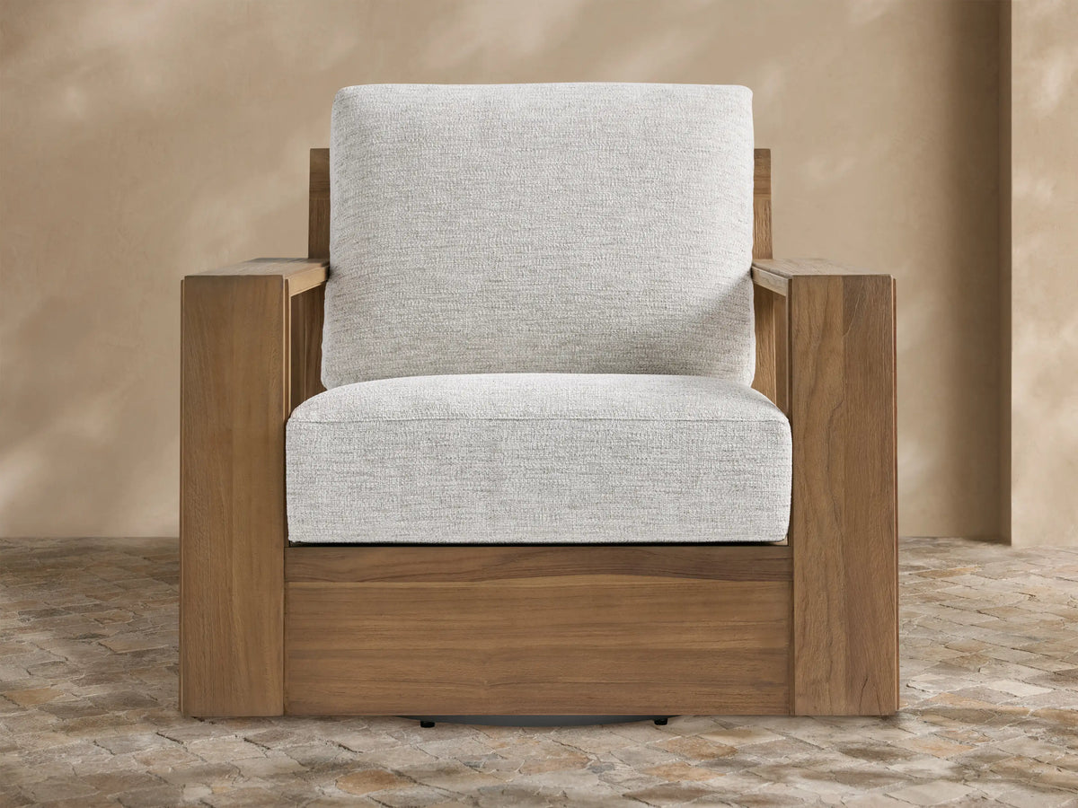 Canyon Outdoor Teak Swivel Chair with golden-brown teak frame & light gray cushions, angled on a rustic tiled patio.