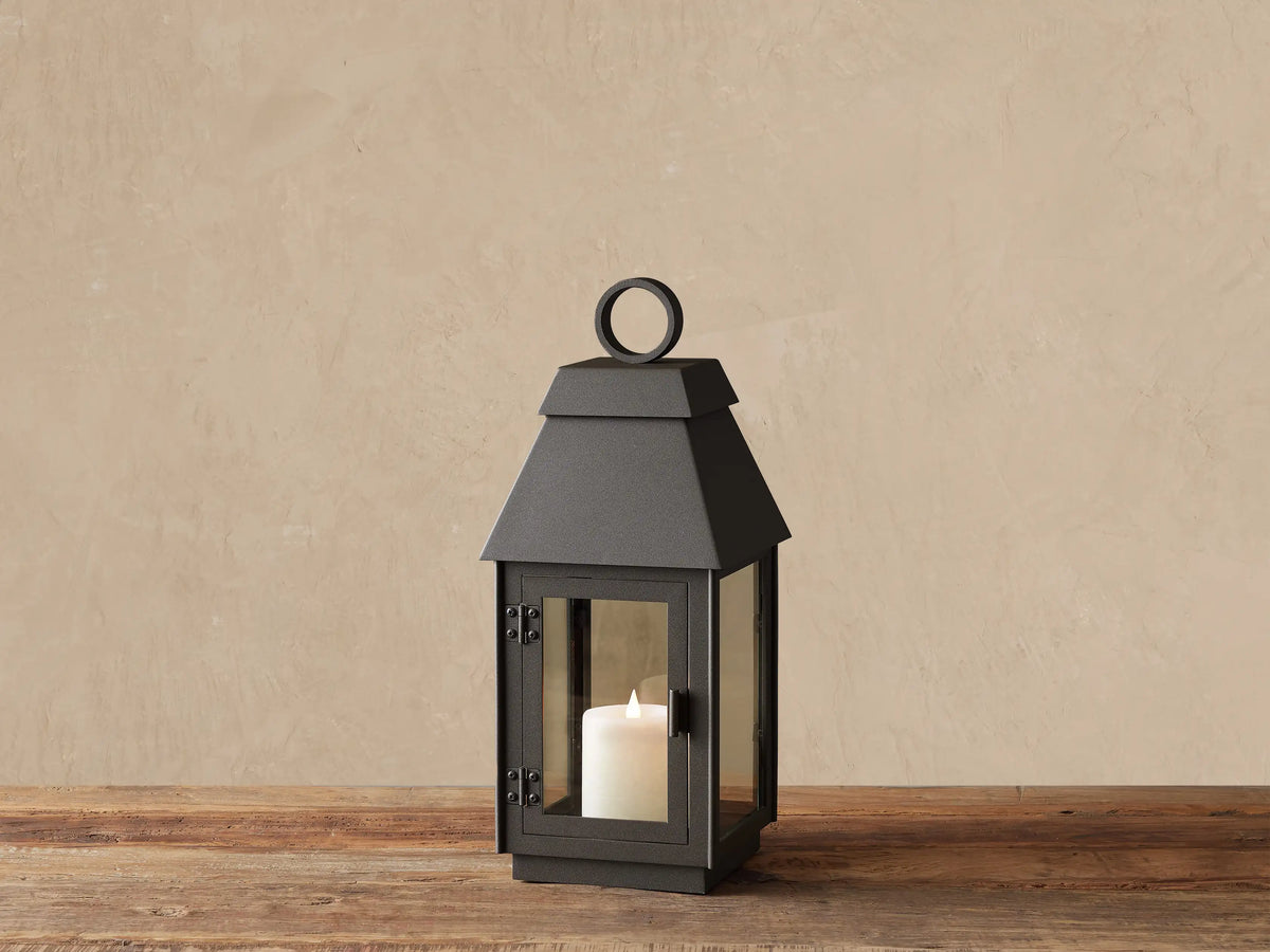 Dark gray Carena Outdoor Lantern on wood. Stainless steel, clear glass sides, sloped roof, ring handle. White flameless candle inside.