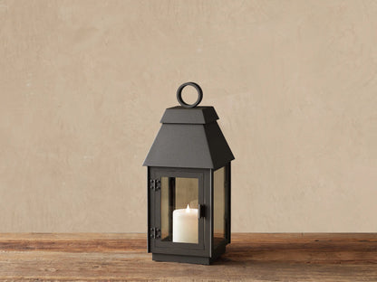 Dark gray Carena Outdoor Lantern on wood. Stainless steel, clear glass sides, sloped roof, ring handle. White flameless candle inside.