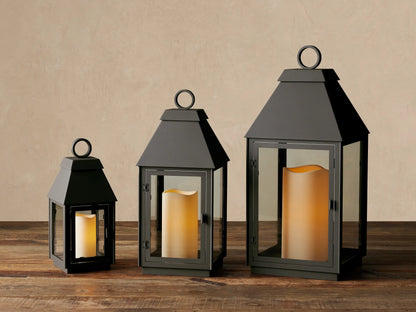 Dark gray Carena Outdoor Lantern on wood. Stainless steel, clear glass sides, sloped roof, ring handle. White flameless candle inside.