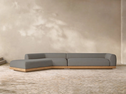 L-shaped Carmel outdoor sectional with gray cushions and a light wooden teak base on a stone patio, with a sunlit wall.