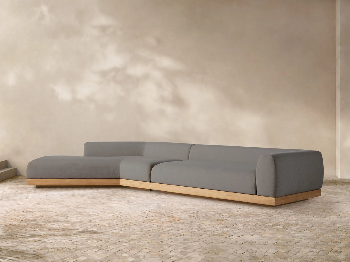 L-shaped Carmel outdoor sectional with gray cushions and a light wooden teak base on a stone patio, with a sunlit wall.