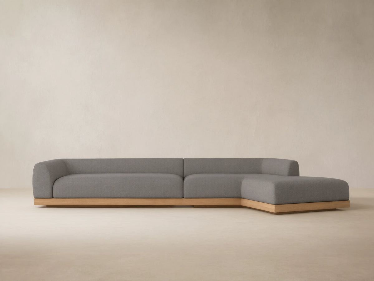 L-shaped Carmel outdoor sectional with gray cushions and a light wooden teak base on a stone patio, with a sunlit wall.