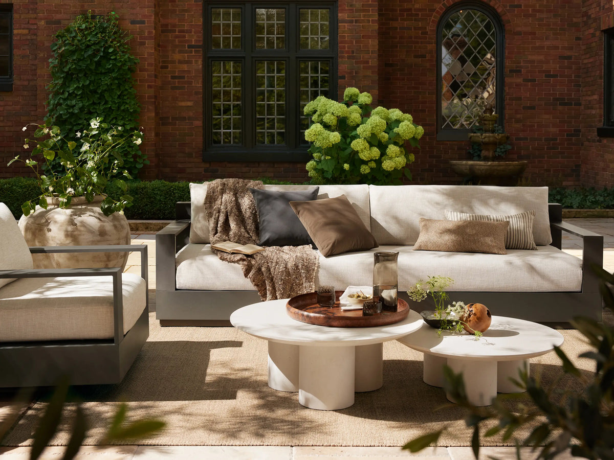 Cascade Outdoor Coffee Table, round, dark brown with a beveled edge, supported by three thick cylindrical legs, on a tiled patio.
