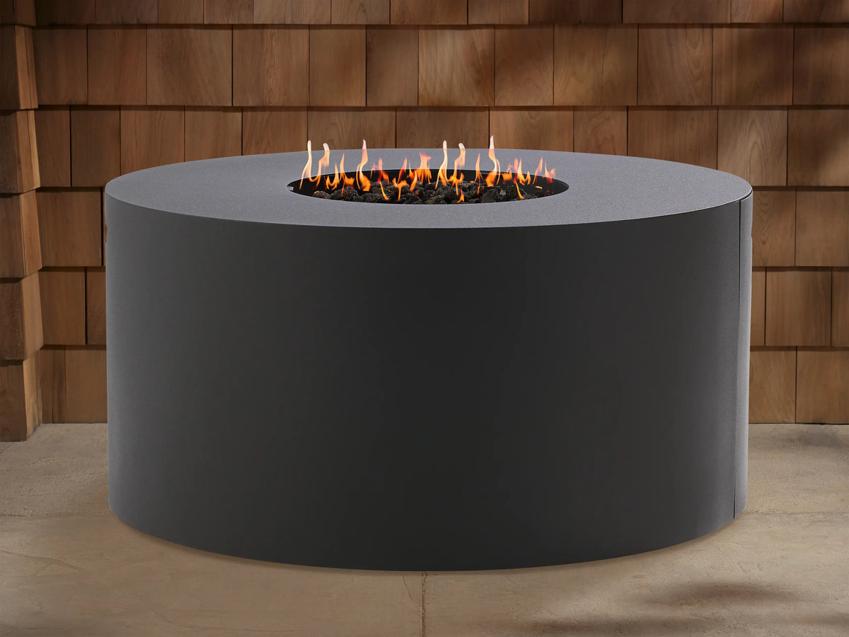 Round, dark gray Coden Outdoor Propane Fire Pit with bright orange and yellow flames burning in the center. Wooden wall in background.