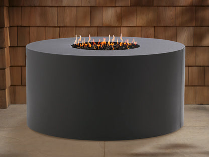 Round, dark gray Coden Outdoor Propane Fire Pit with bright orange and yellow flames burning in the center. Wooden wall in background.