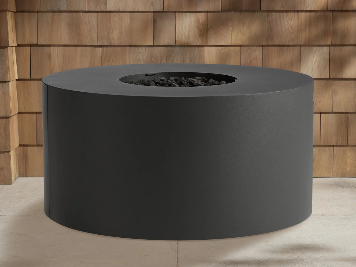 Round, dark gray Coden Outdoor Propane Fire Pit with bright orange and yellow flames burning in the center. Wooden wall in background.