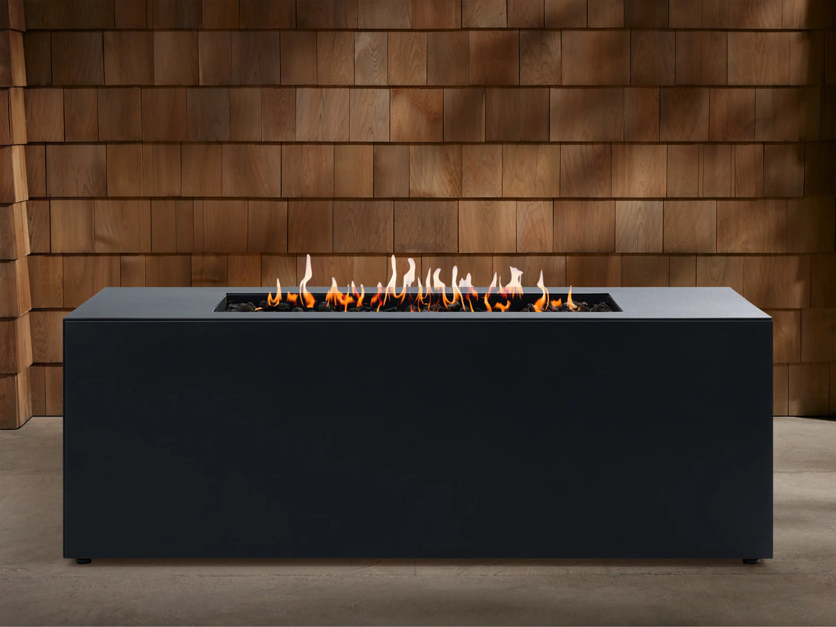 Coden Rectangular Propane Fire Pit in dark grey with orange flames rising from black lava rocks, against a wooden wall.