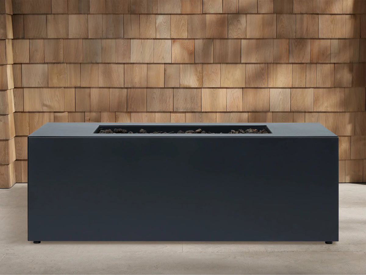 Coden Rectangular Propane Fire Pit in dark grey with orange flames rising from black lava rocks, against a wooden wall.