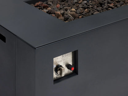 Dark gray rectangular Coden Propane Fire Pit with black lava rocks, shown outdoors on a light patio against a wooden shingle wall.