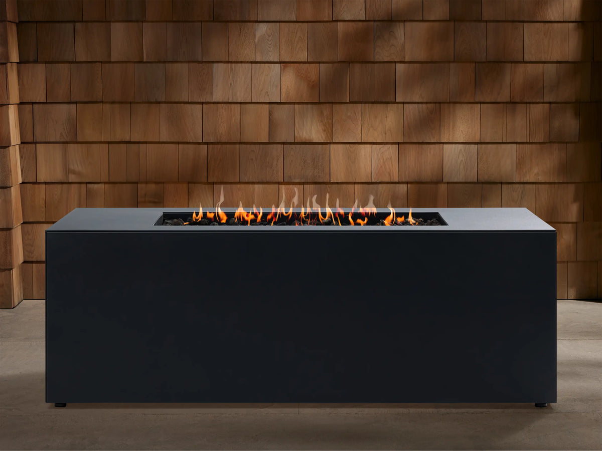 Coden Rectangular Natural Gas Fire Pit, dark gray, with vibrant flames burning in a central trough against a wooden wall.