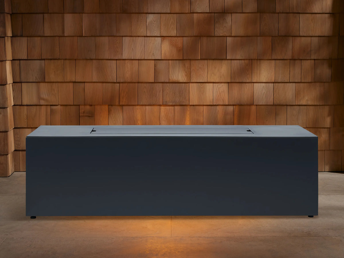 Dark gray Coden Rectangular Propane Fire Pit with flames burning over lava rocks, set against a wooden shingle wall.