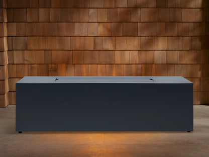 Dark gray Coden Rectangular Propane Fire Pit with flames burning over lava rocks, set against a wooden shingle wall.