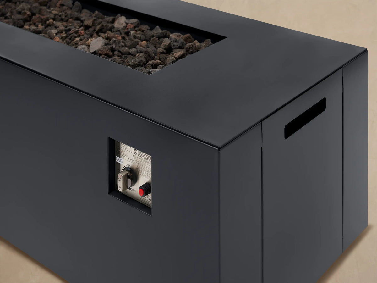 Dark gray Coden Rectangular Propane Fire Pit with lava rocks on a light patio, positioned against a wooden shingle wall.