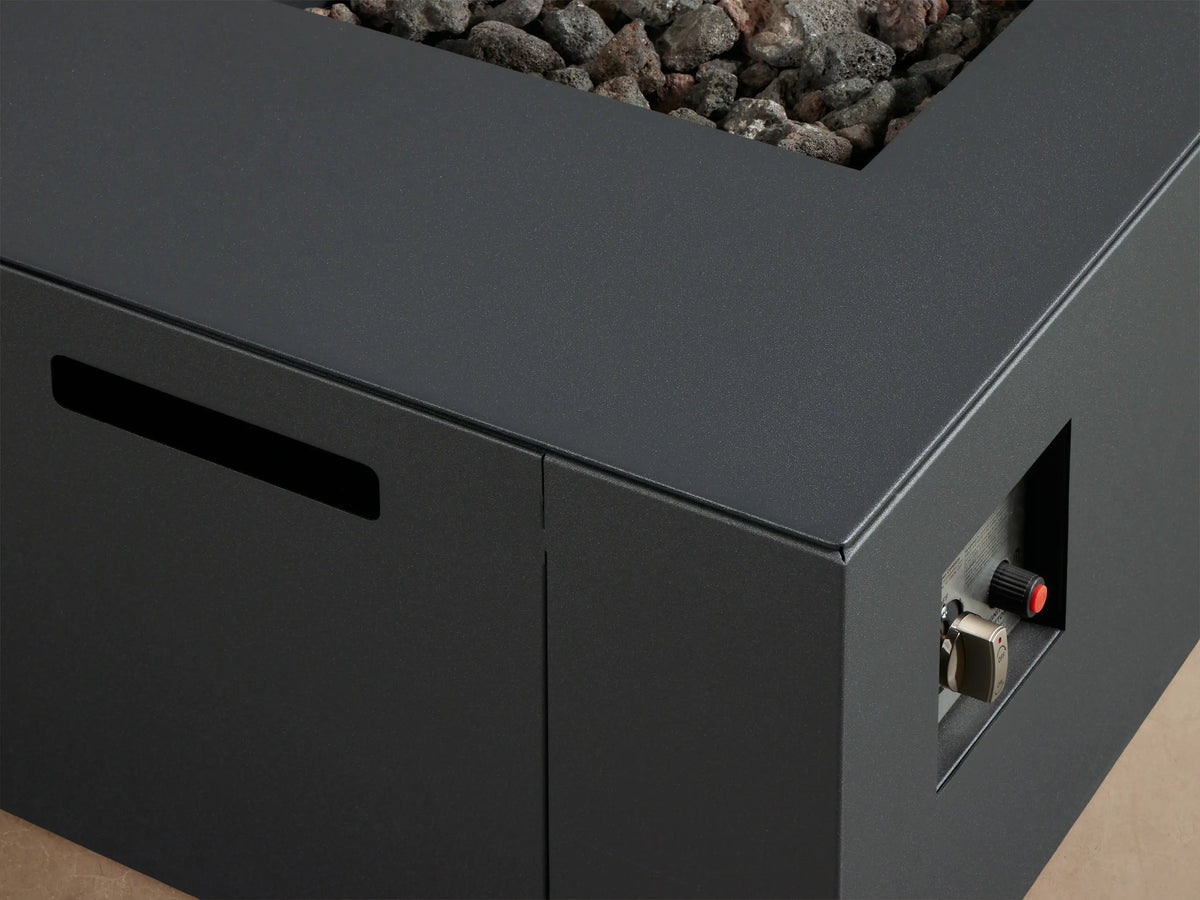Dark gray Coden Rectangular Propane Fire Pit with vibrant orange flames over brown lava rocks, set on a light patio against a wooden wall.
