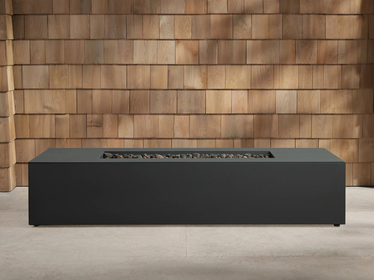 Dark grey Coden Rectangular Propane Fire Pit, centered on a concrete patio with a wooden wall background. Orange flames burn over black lava rocks.