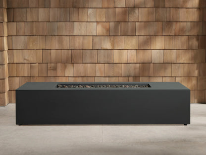 Dark grey Coden Rectangular Propane Fire Pit, centered on a concrete patio with a wooden wall background. Orange flames burn over black lava rocks.