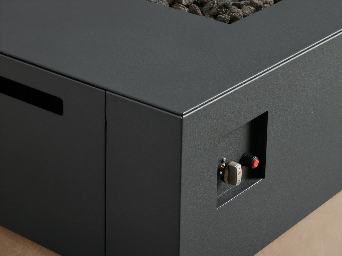 Coden rectangular propane fire pit in dark gray, with lava rocks, on a patio against a natural wooden shingle wall.