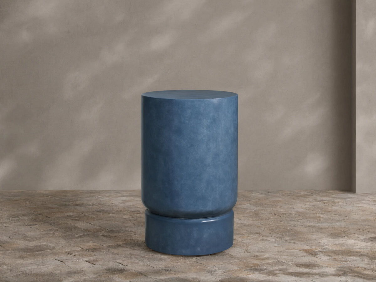 Colorno Outdoor End Table, cylindrical, mottled blue, glossy finish, with recessed base on stone floor against beige wall.