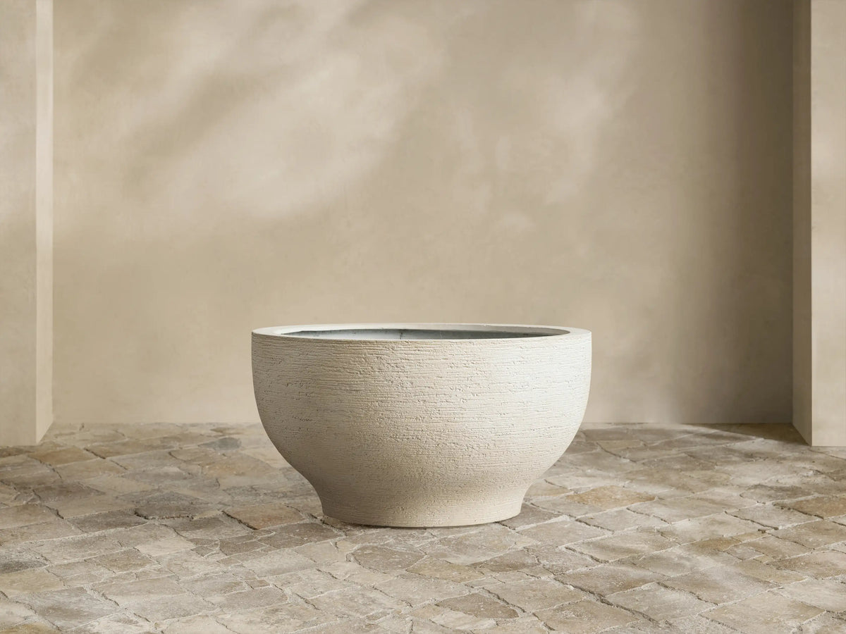 Como Outdoor Planter, round textured pumice-colored, filled with vibrant green variegated plant, on stone tile.