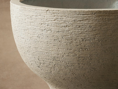 Textured, light pumice-colored, bowl-shaped Como Outdoor Planter on stone pavers against a beige wall.